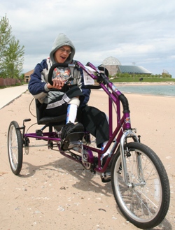 Teen in a sweatshirt rides a purple cycle that has been adapted with three wheels, larger handlebars, and pedal plates with straps